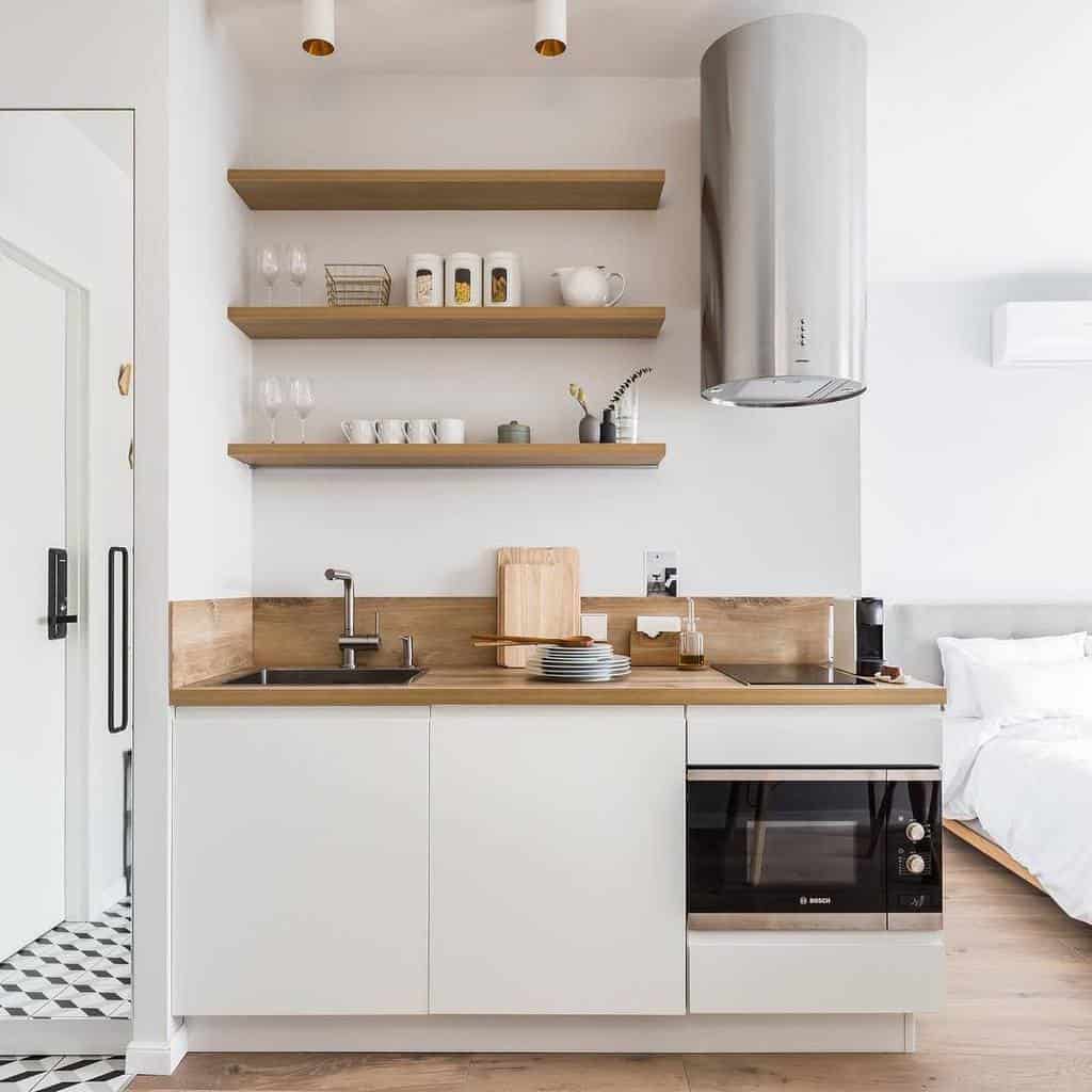 Three wooden floating shelves hold glassware, jars, and decor, adding warmth and function to a sleek, minimalist white kitchen