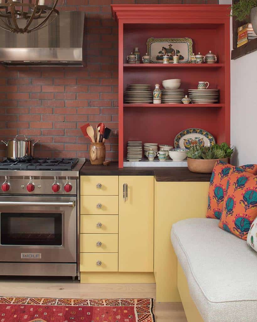 Bold red open shelving showcases decorative plates and ceramics, adding a vintage charm to a colorful kitchen with brick walls and yellow cabinets