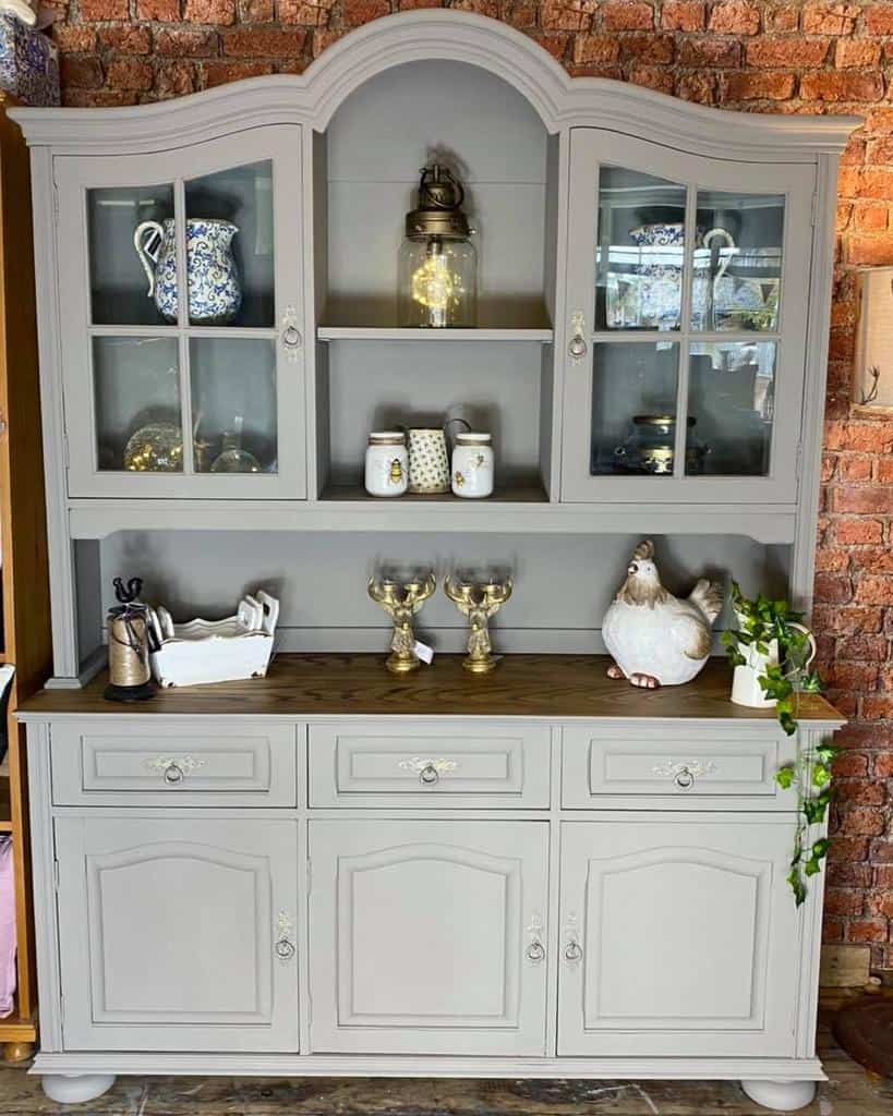 Neutral gray chalk-painted hutch with glass doors, ornate detailing, and a wooden countertop, styled with vintage decor and warm lighting