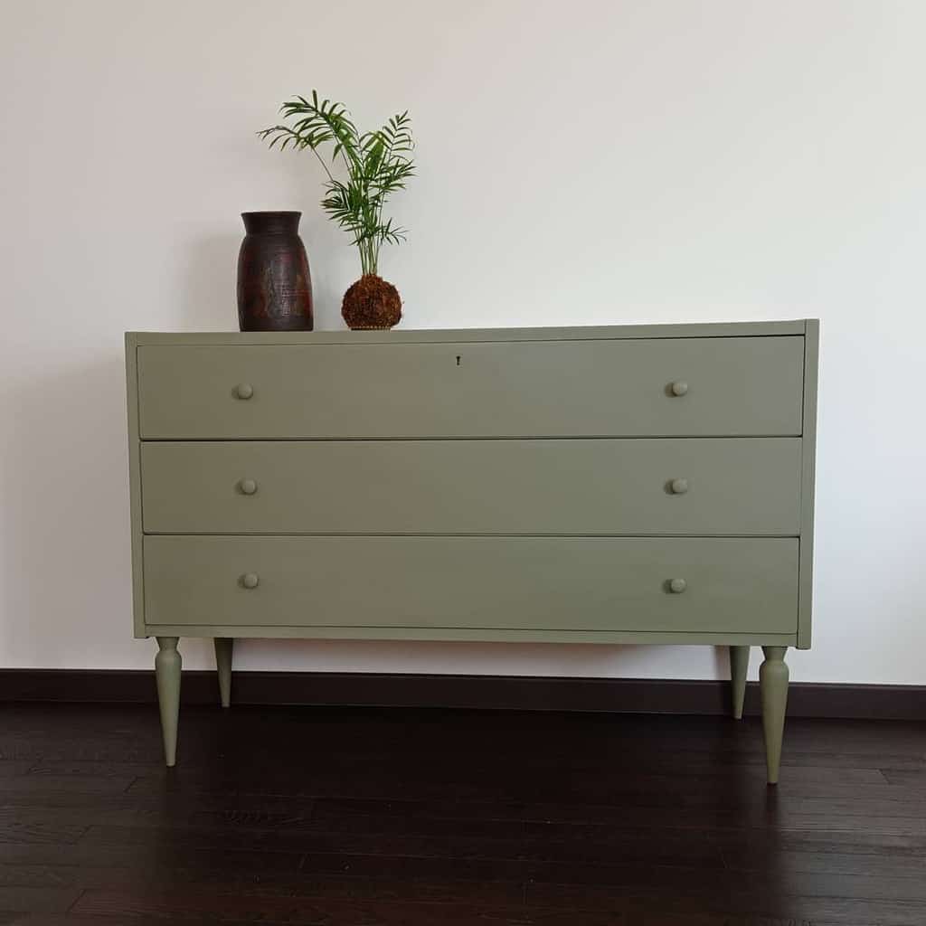 Minimalist sage green chalk-painted dresser with tapered legs and matching knobs, styled with a potted plant and ceramic vase