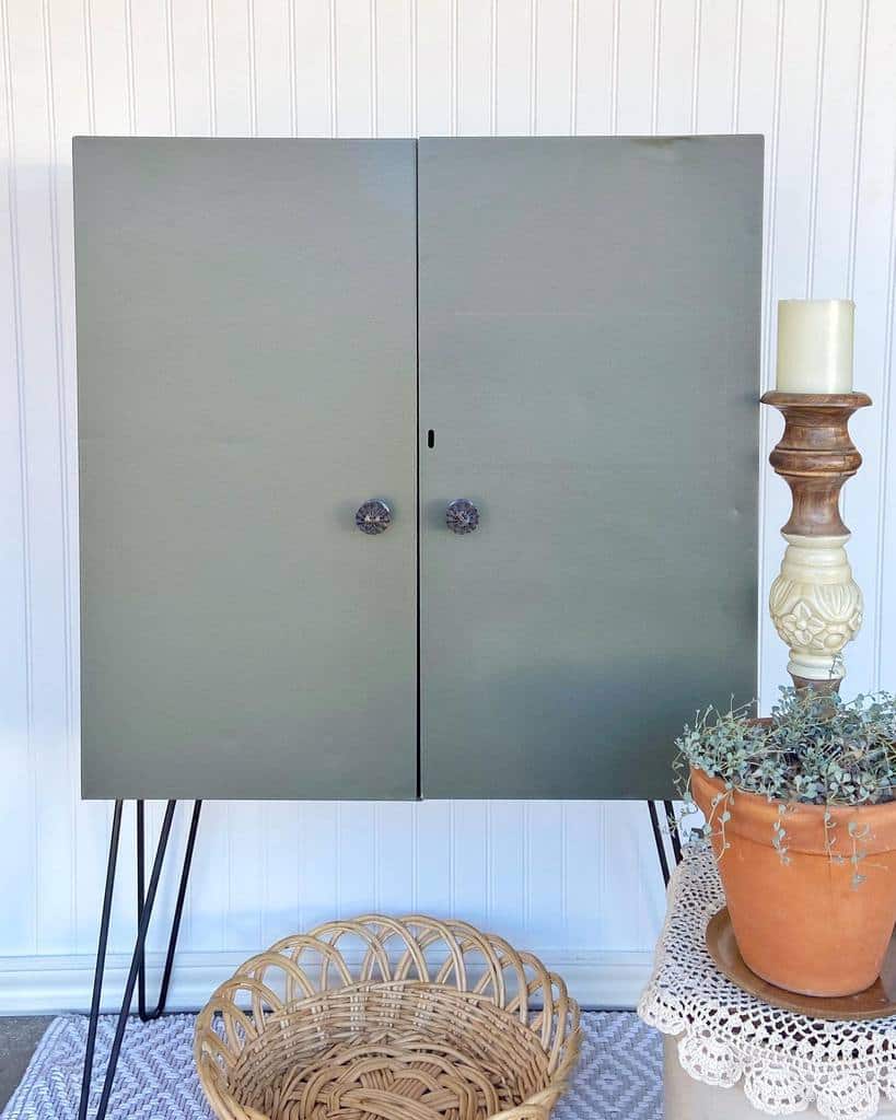 Sage green chalk-painted cabinet with glass knobs and hairpin legs, styled with rustic decor, a wicker basket, and a potted plant