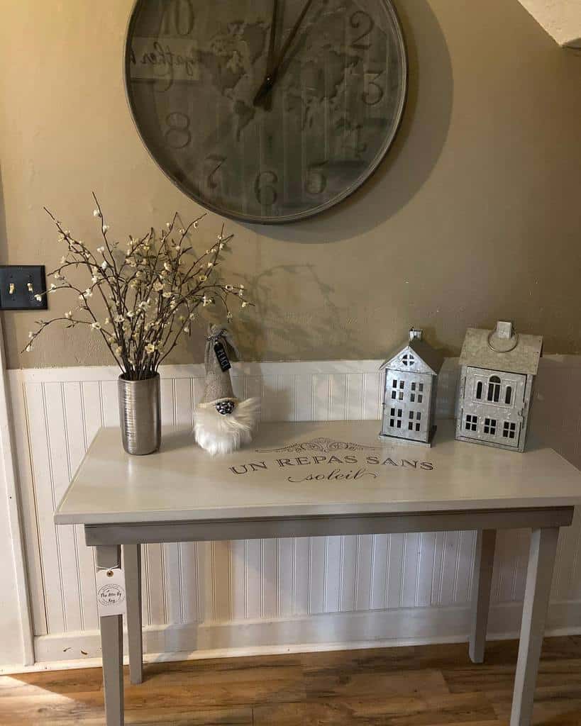 Light gray chalk-painted table with French script design, styled with rustic decor, a large wall clock, and metal house ornaments