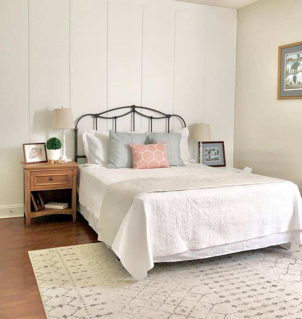 Bedroom with a neatly made bed, white bedding, and a pink throw pillow, wooden side table with lamps and a rug on the floor