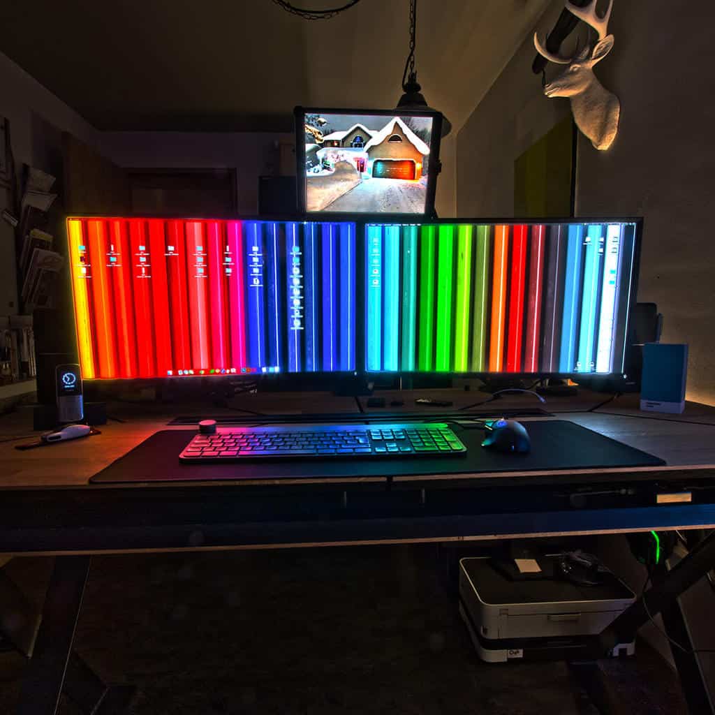 A dual-monitor setup with colorful striped screens on a desk, keyboard, mouse, overhead shelf, and deer head wall decor