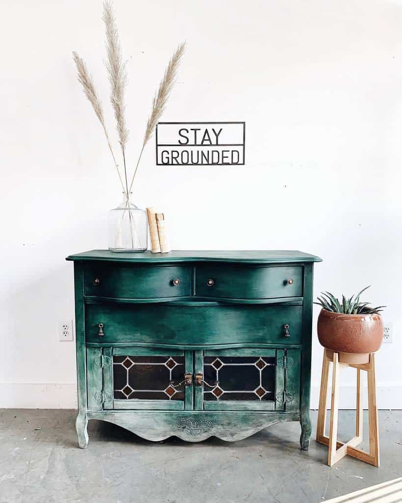 Vintage sideboard with an ombre green chalk-painted finish, stained glass cabinet doors, and rustic decor, set against a minimalist white wall