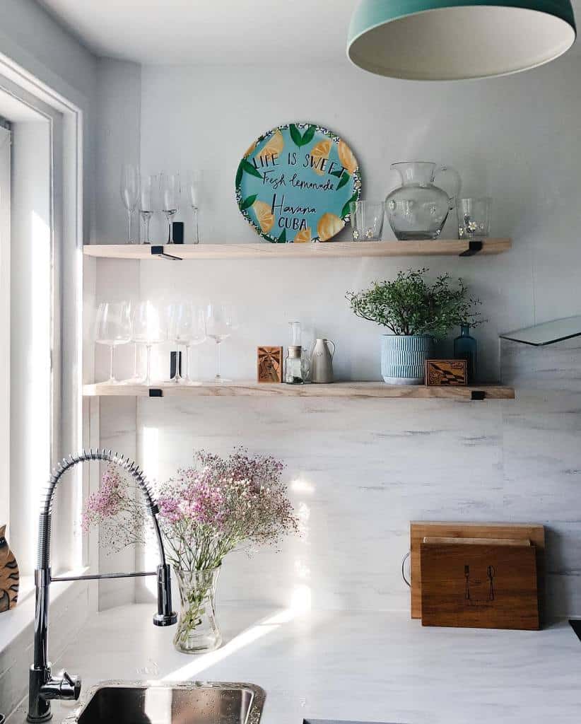 Bright kitchen corner with a sink, flowers in a vase, and wooden shelves holding glassware and a decorative plate