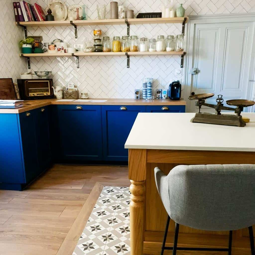 Rustic wooden shelves with black metal brackets display glass jars, ceramics, and books, complementing the farmhouse-style kitchen with blue cabinets