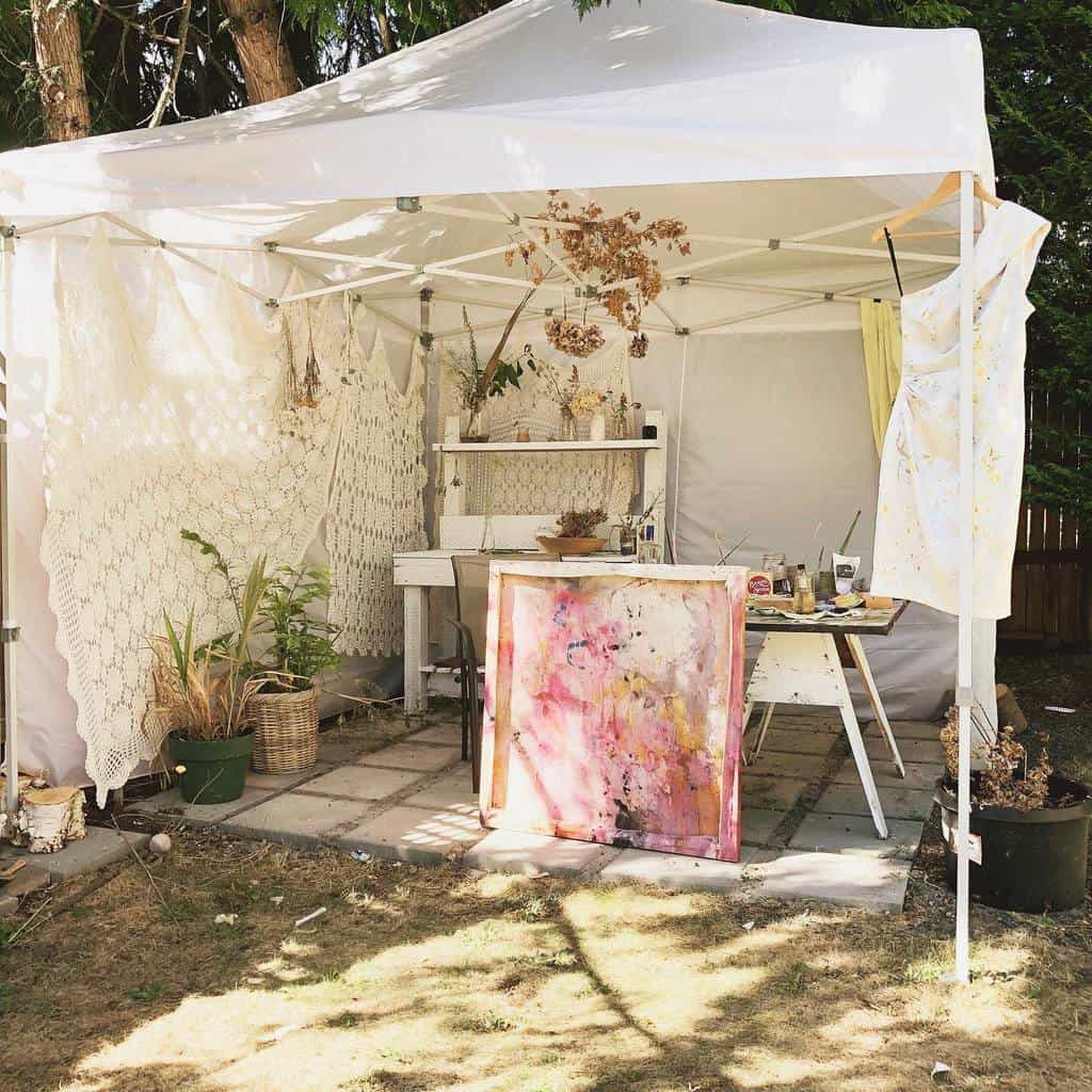 Outdoor art studio under a white canopy with a table, paints, and a vibrant pink and yellow canvas, surrounded by plants and lace curtains