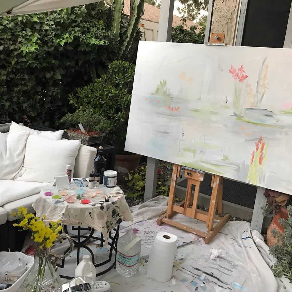 Outdoor painting setup with abstract art on easel, surrounded by art supplies and cozy seating, lush greenery in the background