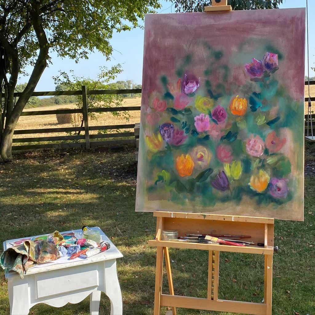 A colorful floral painting on an easel outdoors, with a paint palette and brushes on a small table nearby