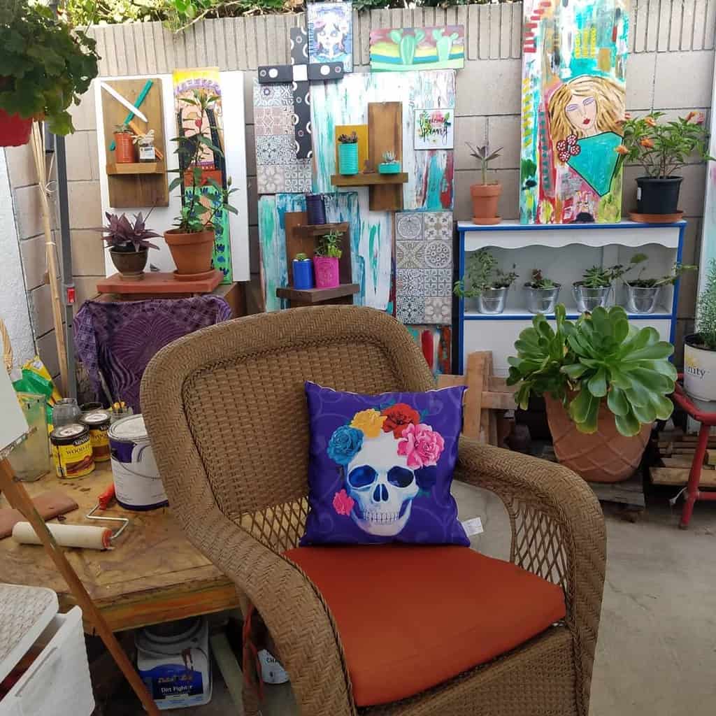 A wicker chair with a colorful skull pillow sits in an eclectic garden; art, plants, and tools are displayed on a wall in the background