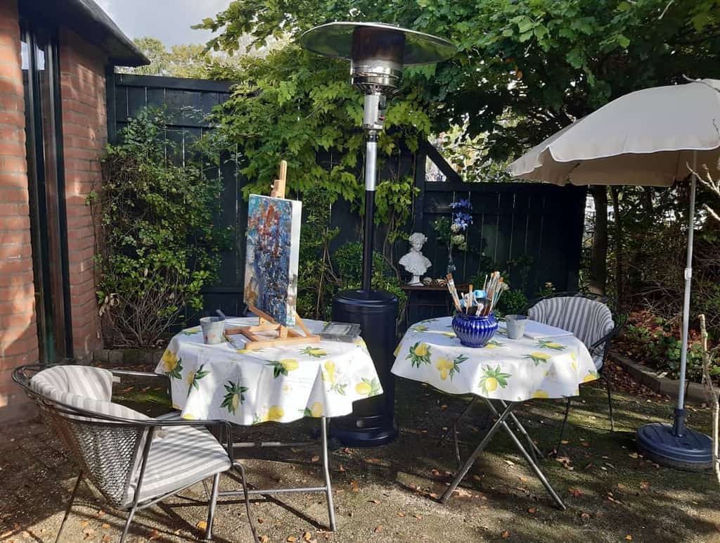 Outdoor art setup: two tables with lemon-patterned tablecloths, an easel with a painting, chairs, a heater, umbrella, and a garden statue