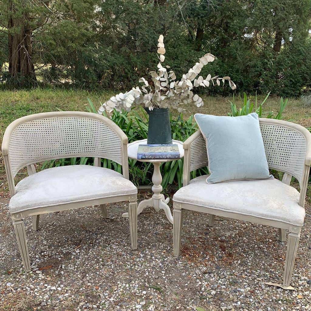 Pair of vintage cane-back chairs with chalk-painted fabric and a distressed neutral frame, styled with a side table and soft decor outdoors