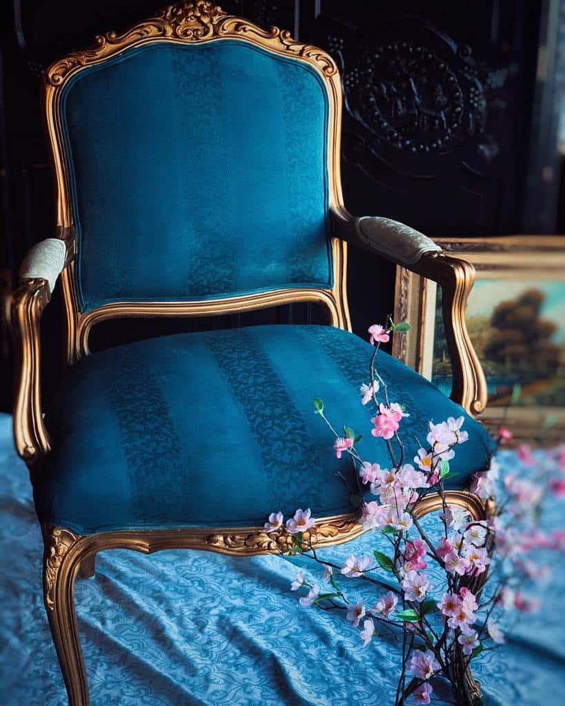 Luxurious vintage armchair with deep teal chalk-painted fabric and an ornate gold frame, styled with cherry blossoms and antique decor