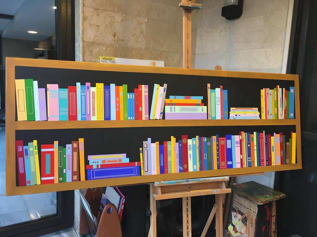 Colorful, minimalist painting of books on two shelves, displayed on an easel in a room with stone walls