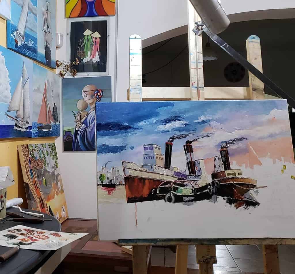 Art studio with an easel displaying a work-in-progress painting of ships, surrounded by nautical and portrait artworks on the walls