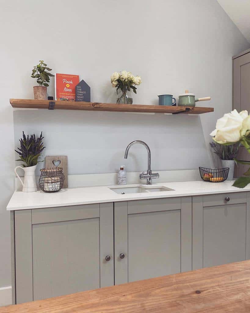 A single rustic wooden shelf with black brackets holds plants, books, and kitchen decor, complementing a minimalist farmhouse-style sink area