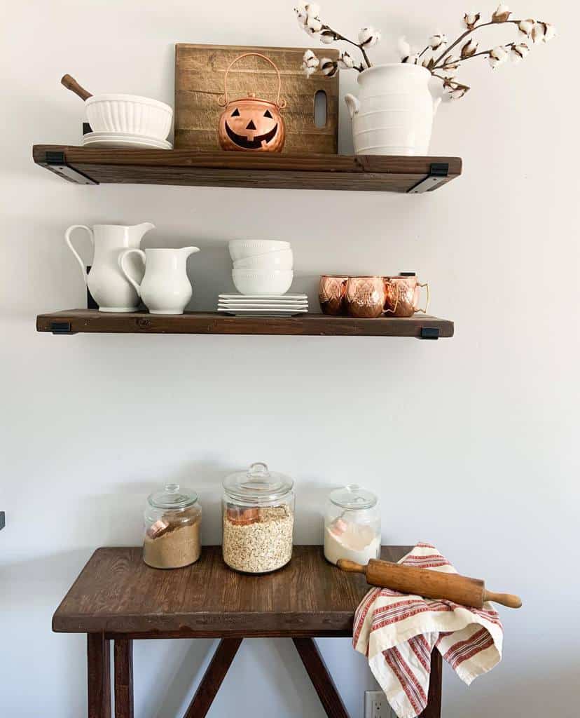 Kitchen decor with shelves holding white dishes, copper mugs, a pumpkin lantern, jars with ingredients, and a rolling pin on a towel