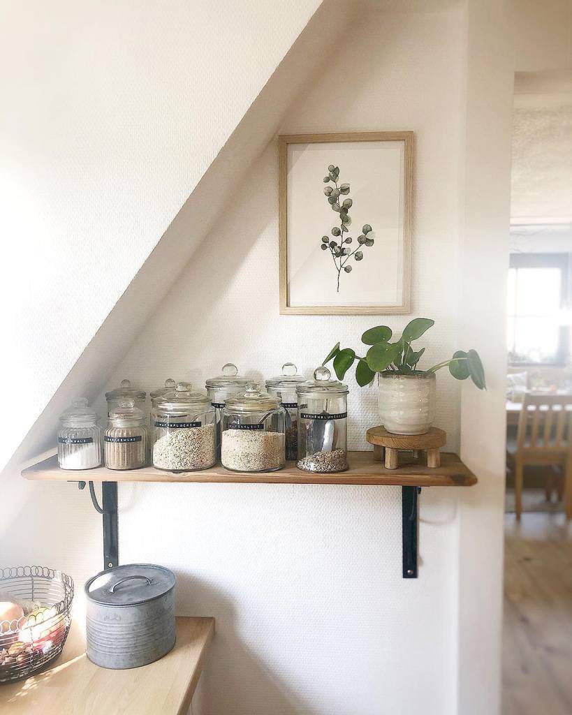 Kitchen corner with a shelf holding jars of grains and a plant, framed botanical print above, dining area visible in the background