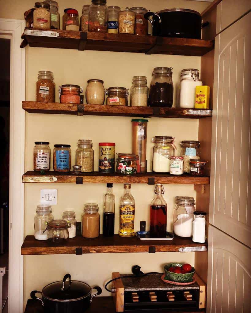 Wooden kitchen shelves with jars of spices, grains, and cooking essentials; a small table holds bottles and a bowl of tomatoes