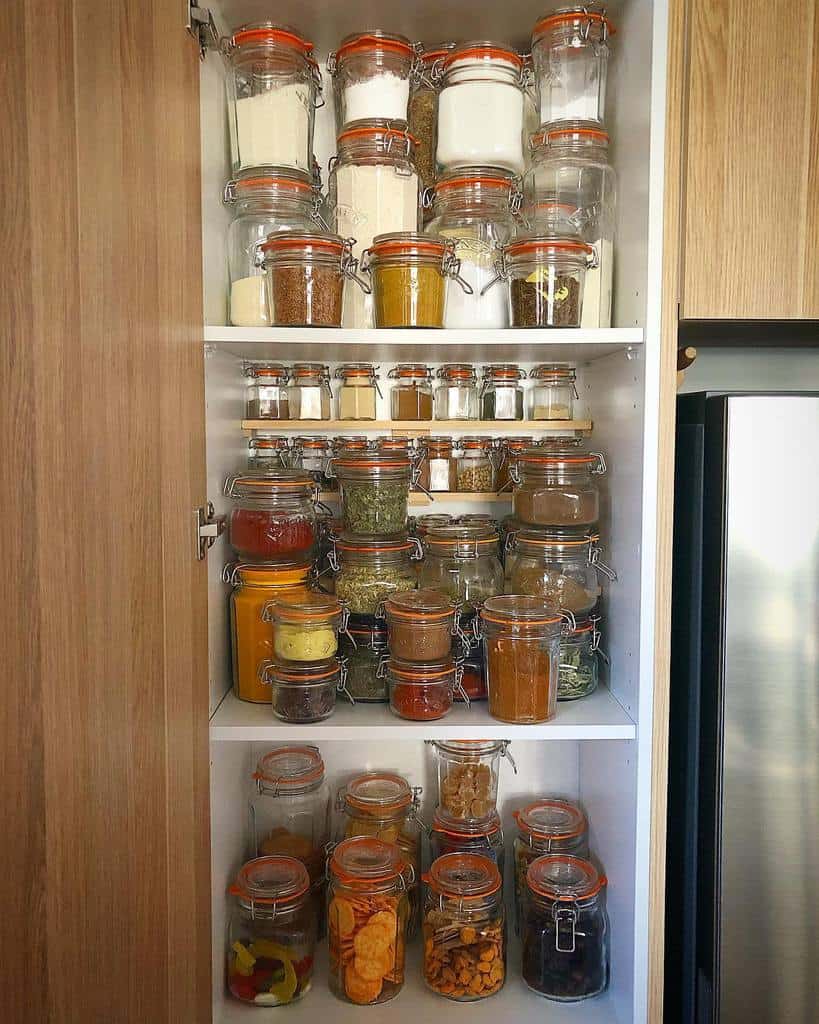 A pantry shelf filled with various sealed jars containing spices, grains, and dried foods, neatly organized in a wooden cabinet