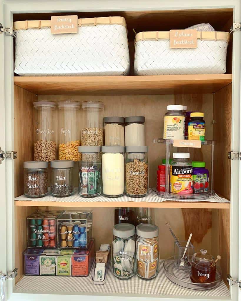 Organized pantry shelves with labeled baskets, jars of cereals and grains, vitamin bottles, teas, honey, and kitchen essentials