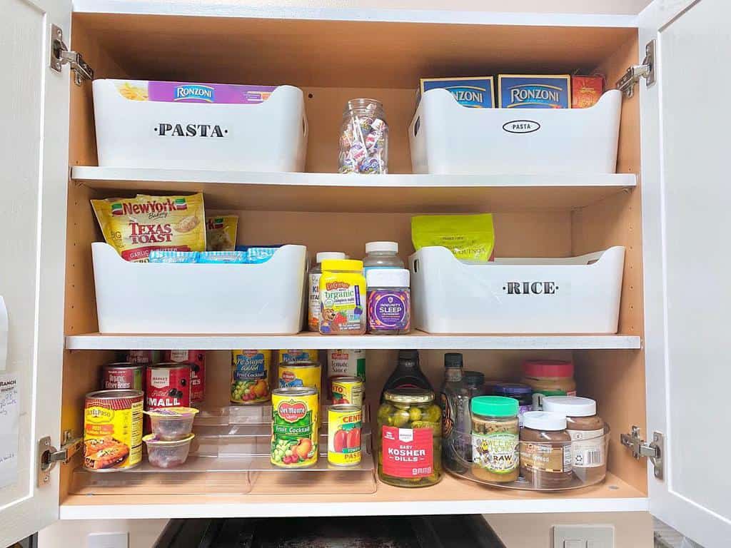 Kitchen cupboard organized with labeled containers for pasta and rice, assorted snacks, canned goods, and spice jars