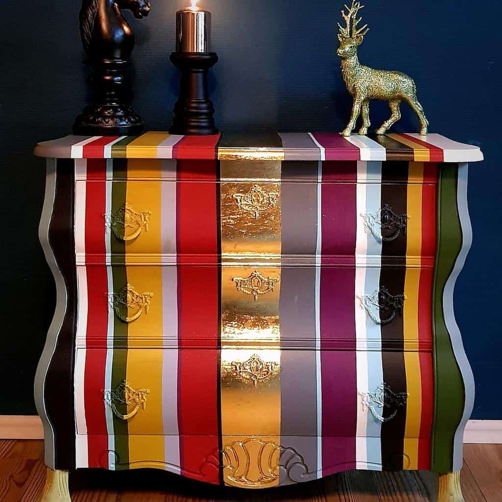 Bold striped chalk-painted dresser with gold leaf accents, curved edges, and ornate hardware, styled with dramatic dark decor