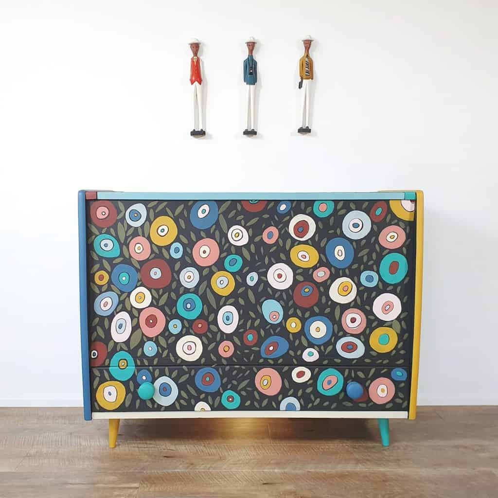 Colorful floral-patterned chalk-painted dresser with mid-century modern legs, set against a white wall with quirky wooden figurines