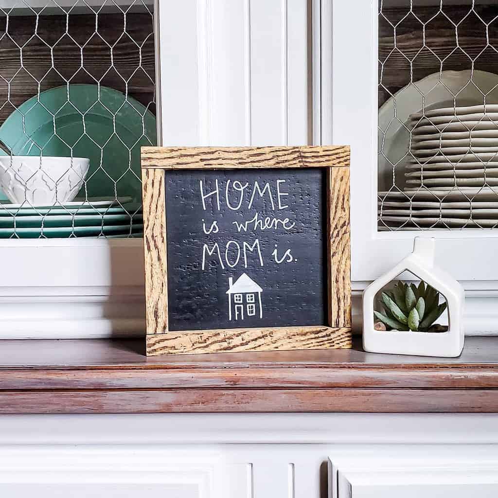 Decorative sign reading "Home is where mom is" beside a small plant on a shelf with dishes in the background