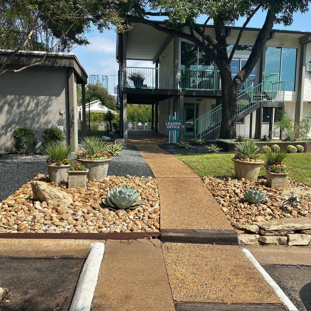 Modern xeriscape entrance with rock gardens, succulents, and potted plants leading to a leasing office with a tree-lined walkway