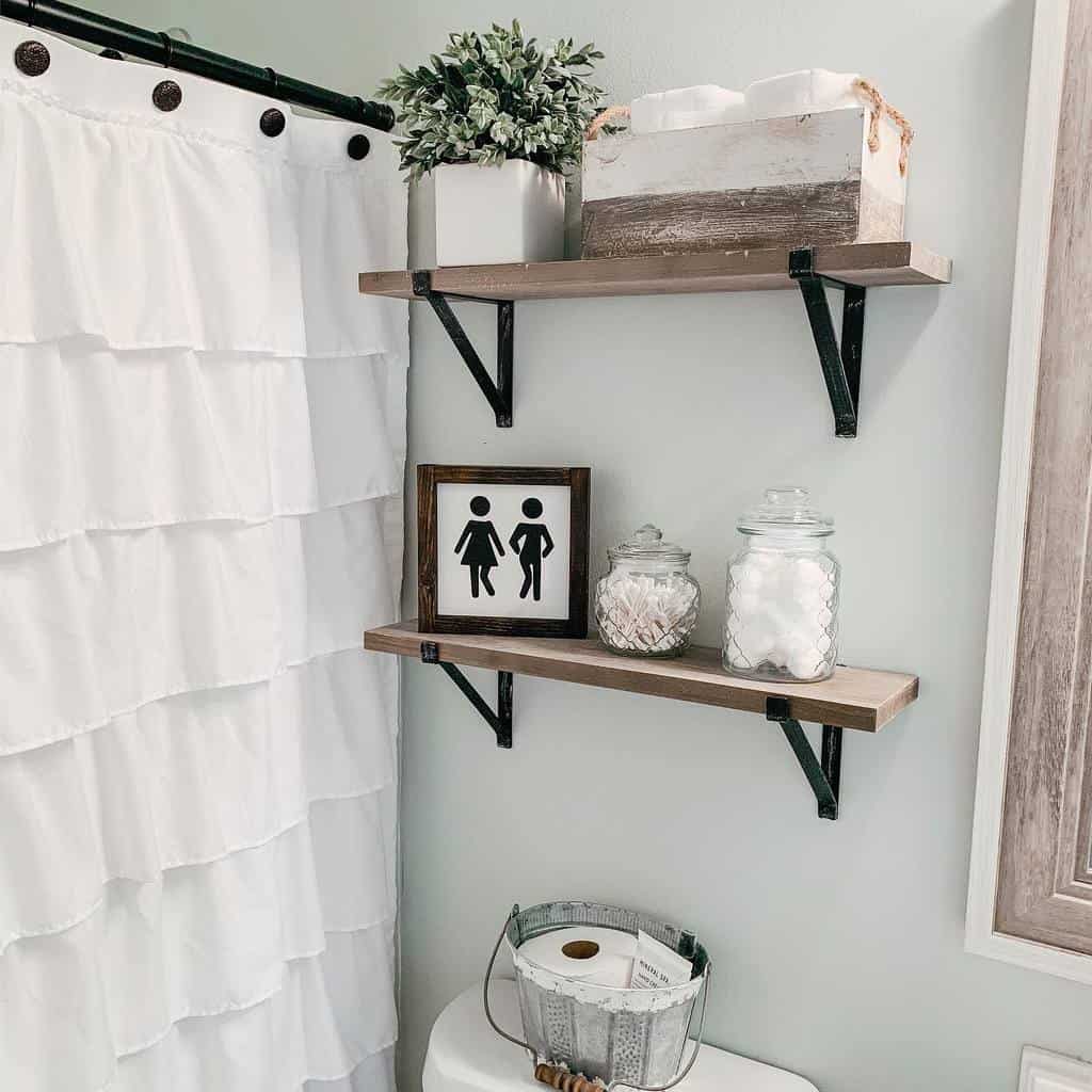 Bathroom corner with ruffled shower curtain, two wooden shelves holding a plant, jars, sign, and a tissue box above a toilet