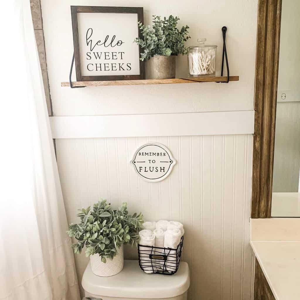 Bathroom decor with plants, "Hello Sweet Cheeks" sign on shelf, and "Remember to Flush" sign above toilet with a basket of toilet paper