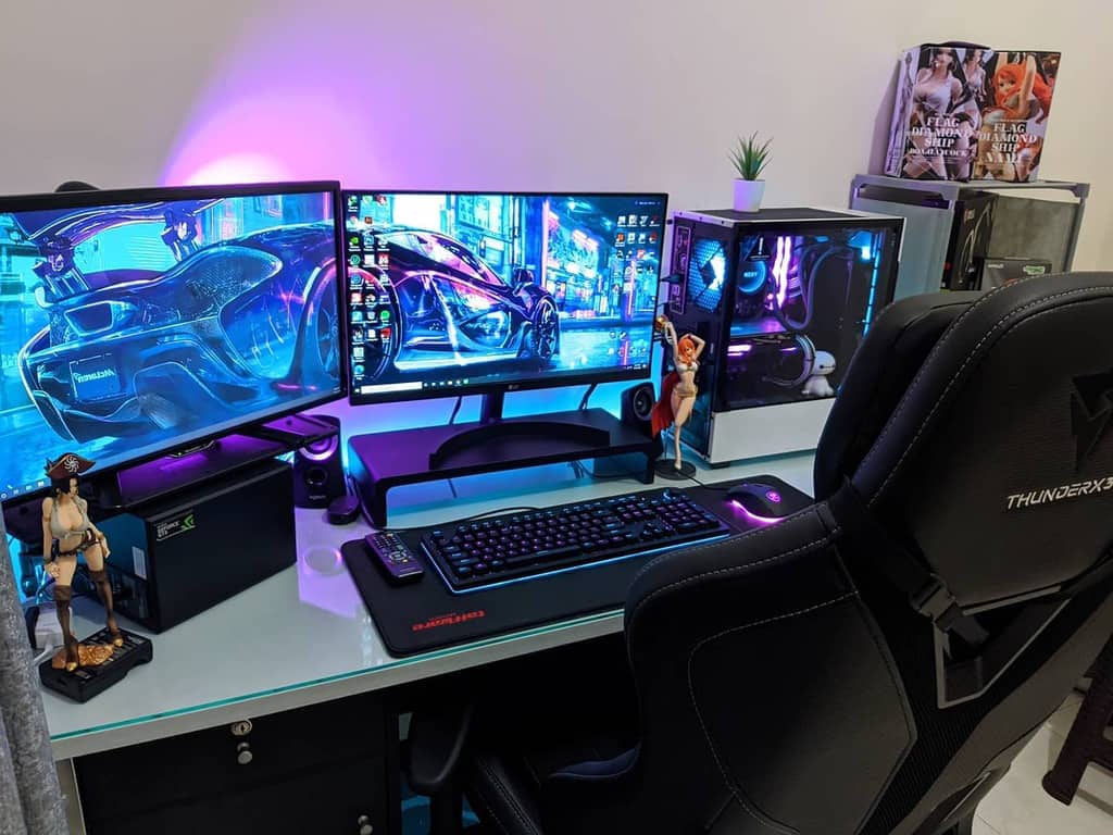 Gaming setup with dual monitors displaying futuristic car art, RGB-lit PC, gaming chair, keyboard, mouse, and action figures on desk