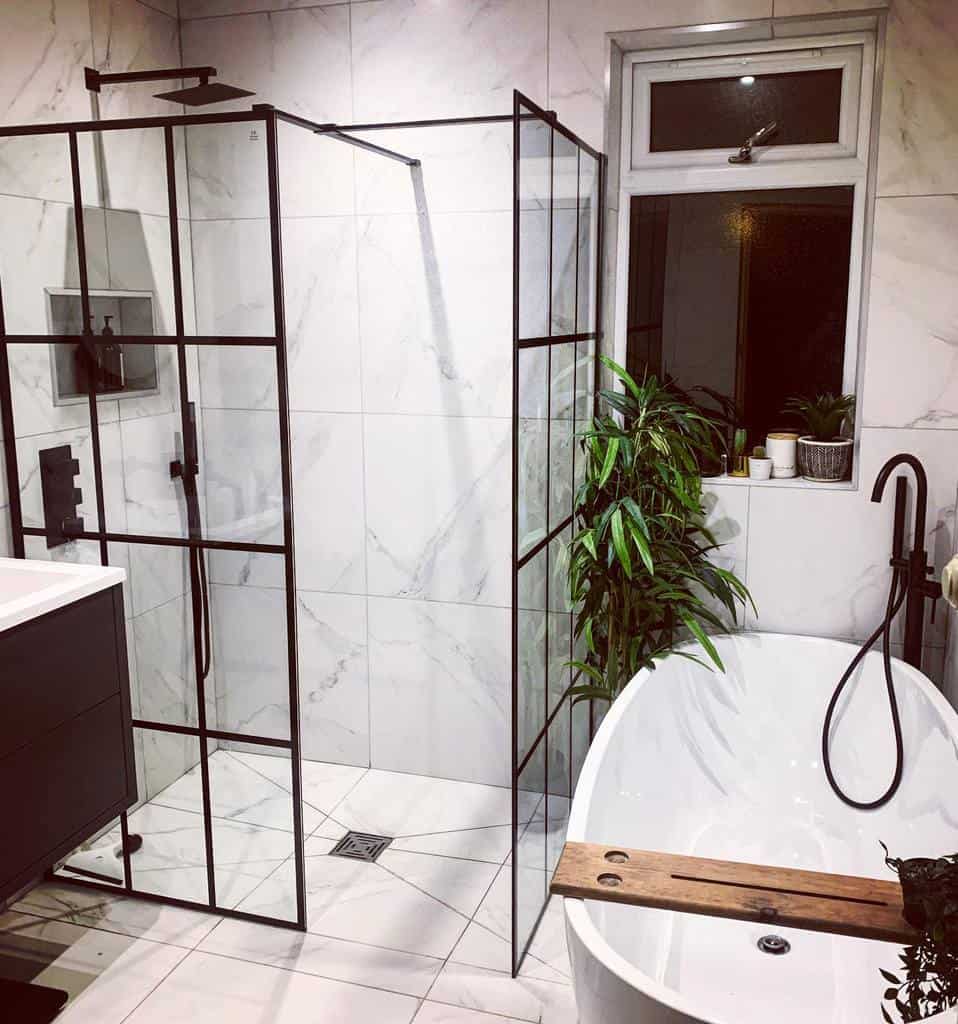 Modern wet room with a black-framed glass shower, white freestanding bathtub, potted plants, and marble tile walls and floor