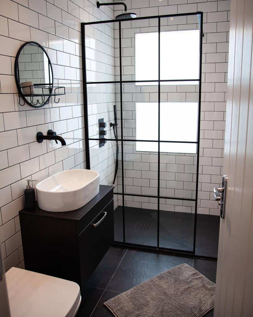 Modern wet room with glass shower, white subway tiles, black fixtures, and a round mirror; a dark vanity holds a sleek vessel sink