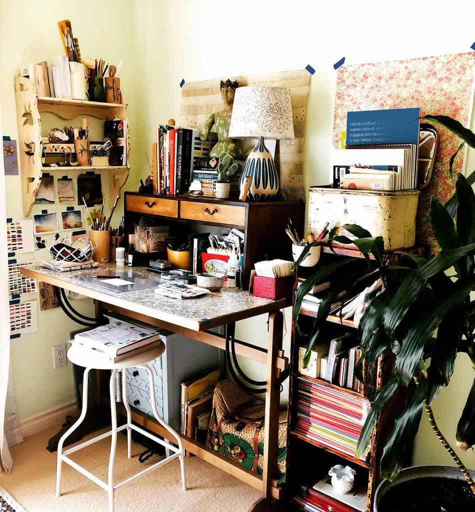 Cozy art studio corner with wooden desk, shelves of art supplies, books, plants, and a patterned chair