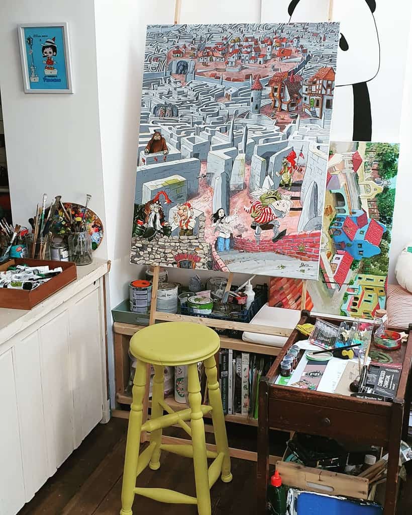 Art studio with a colorful painting on an easel, surrounded by art supplies, a yellow stool, and a cluttered workspace