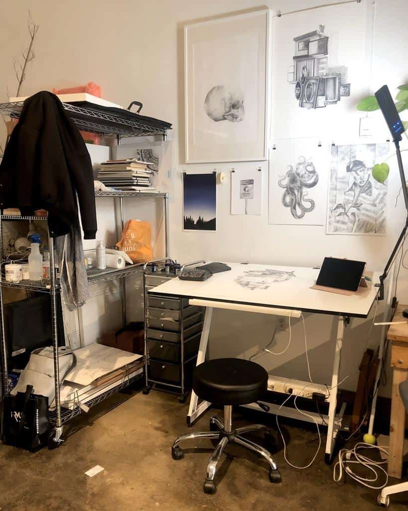 Art studio corner with a desk, sketches on the wall, a stool, and storage shelves with art supplies and a jacket