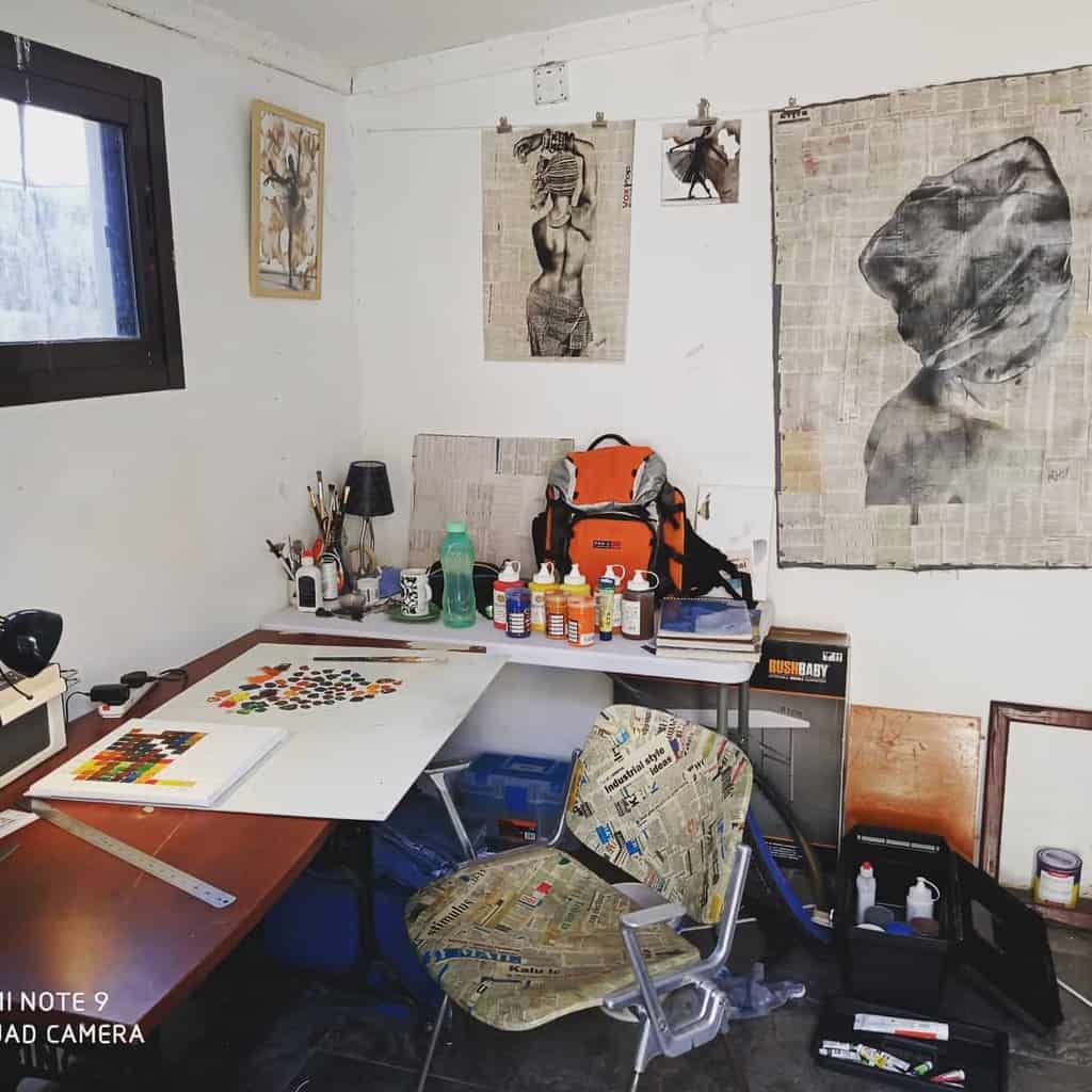 Art studio with a desk, art supplies, and a collage-covered chair; walls display abstract artworks on newspaper backgrounds