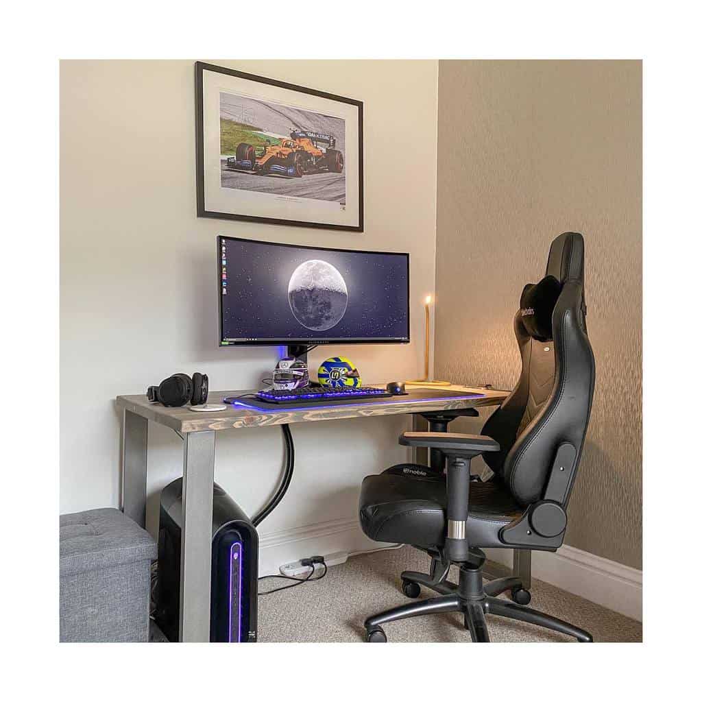 A modern desk setup with a large monitor displaying a moon image, gaming chair, keyboard, mouse, headphones, and a racing car picture above