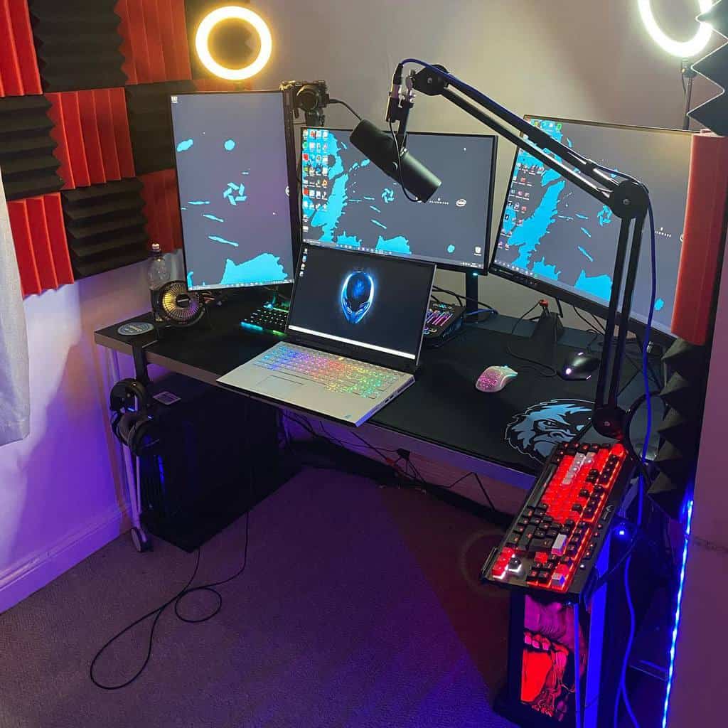 Gaming setup with a laptop, three monitors, gaming keyboard, mouse, microphone, two ring lights, and red acoustic foam on the wall