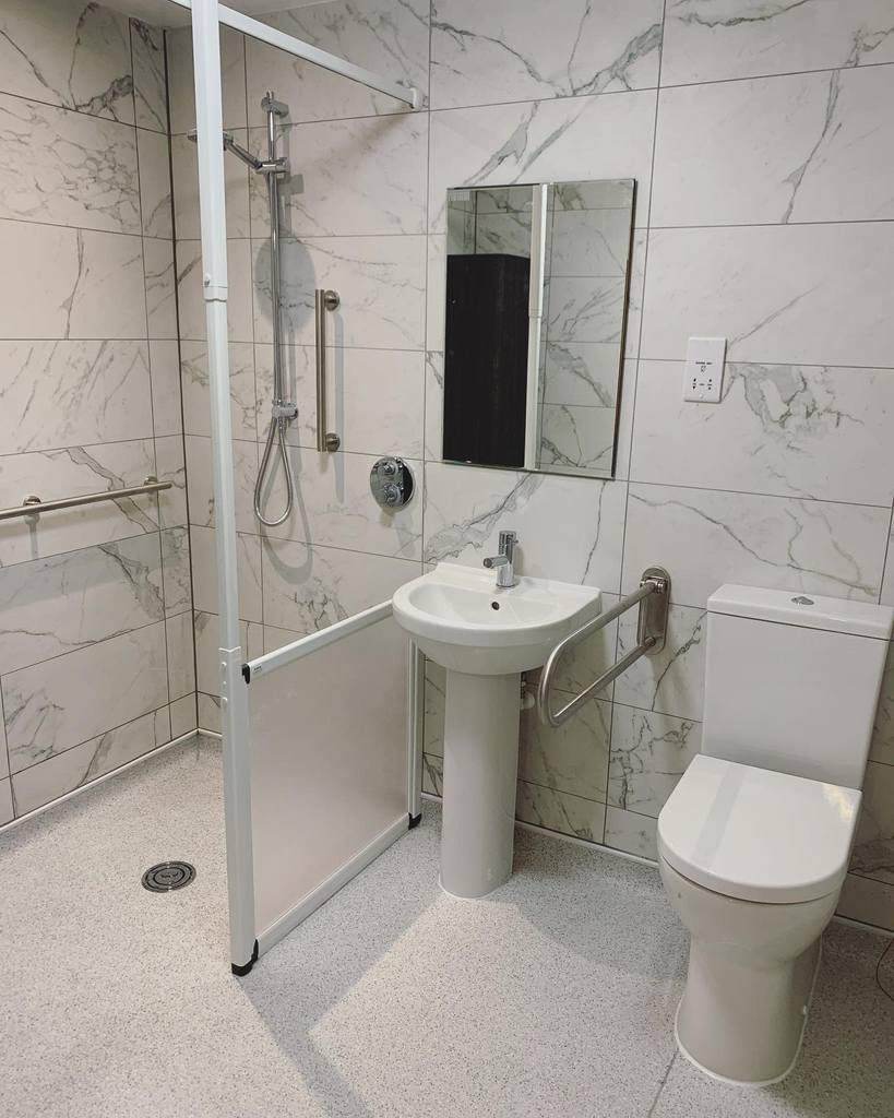 Accessible wet room with a toilet, sink, mirror, and walk-in shower featuring grab bars and elegant marble-patterned tiles