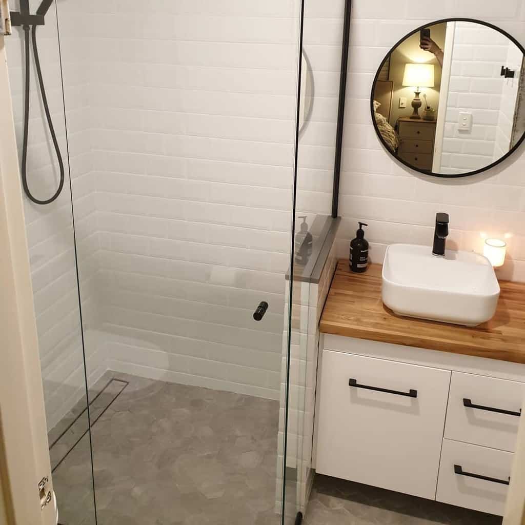 Minimalist wet room with white tiles, glass shower, wooden countertop, round mirror, and modern sink; soft lighting creates a cozy ambiance