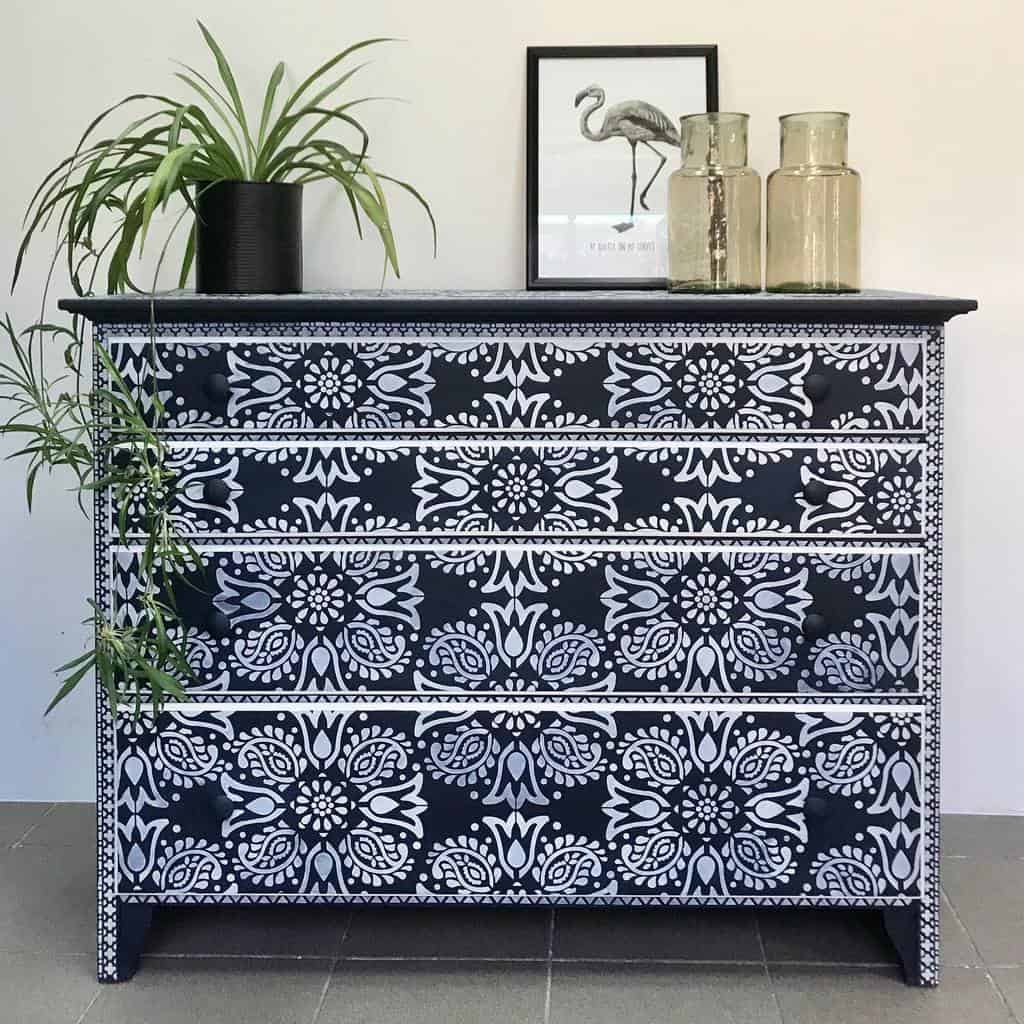 Black chalk-painted dresser with intricate white stencil patterns, styled with greenery, glass vases, and a framed flamingo print