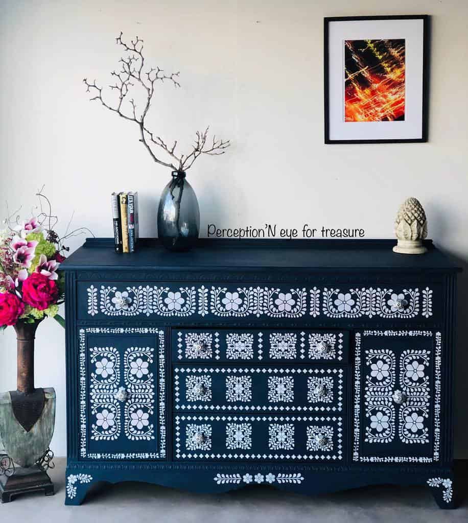 Dark blue chalk-painted dresser with intricate white floral stencil patterns, styled with modern decor, a vase with branches, and bold artwork