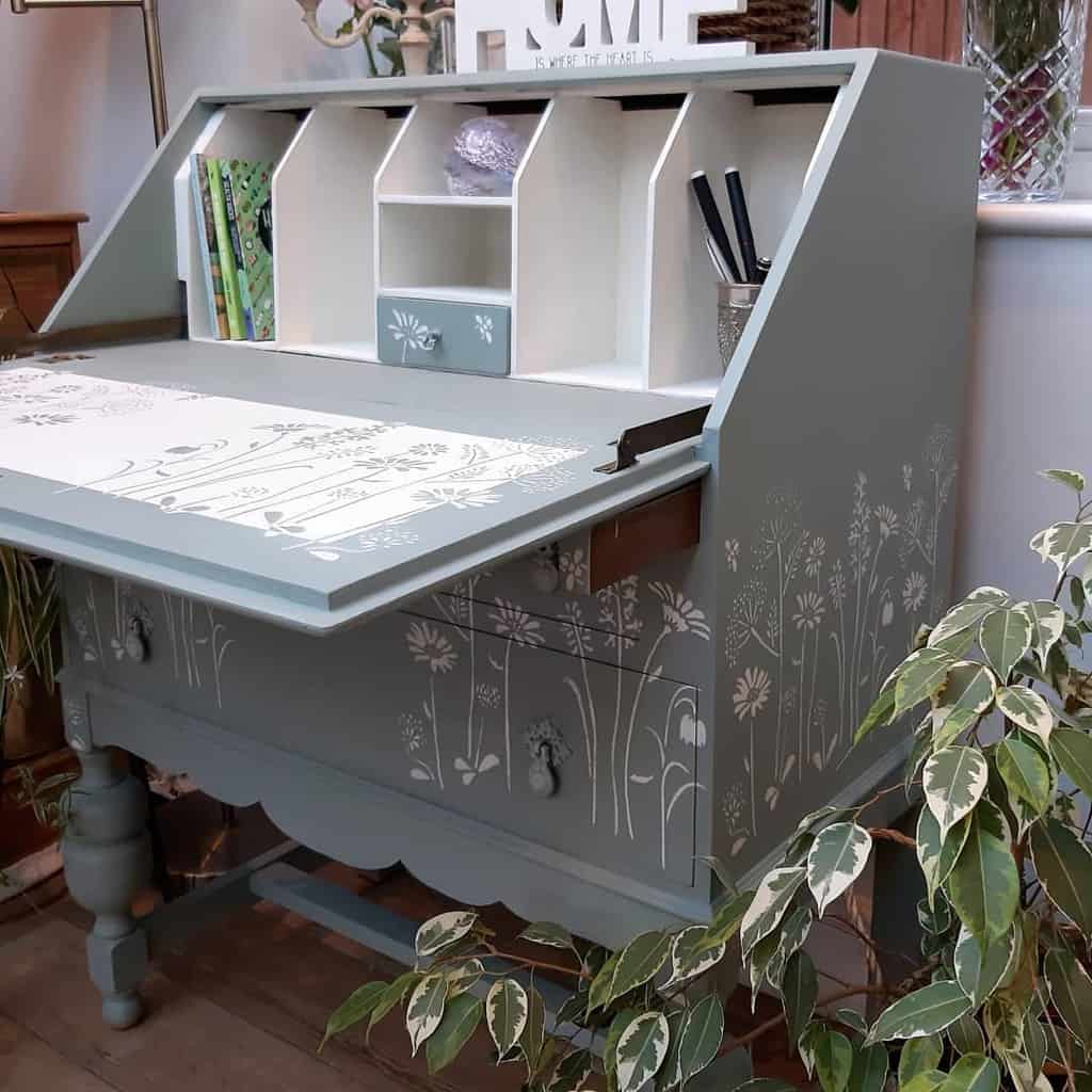 Vintage secretary desk painted in muted green chalk paint with delicate floral stencils, featuring organized storage and rustic charm