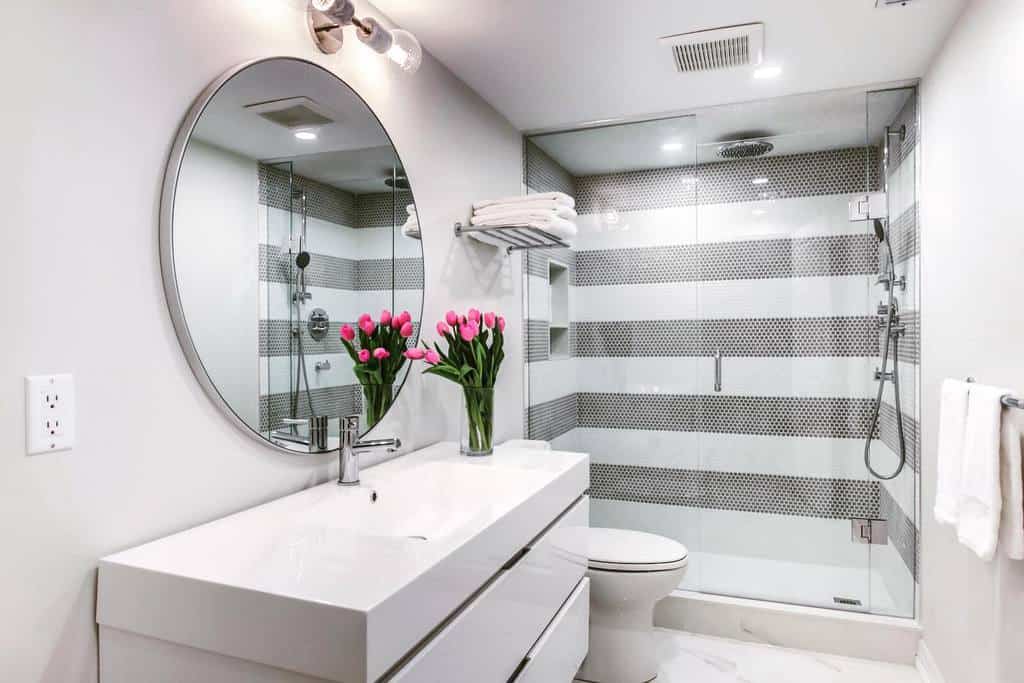 Modern wet room with a round mirror, white vanity, vase of pink tulips, and glass shower featuring striped tiles