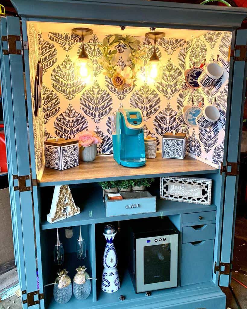 Upcycled armoire transformed into a stylish coffee bar with blue chalk paint, patterned wallpaper, warm lighting, and organized storage