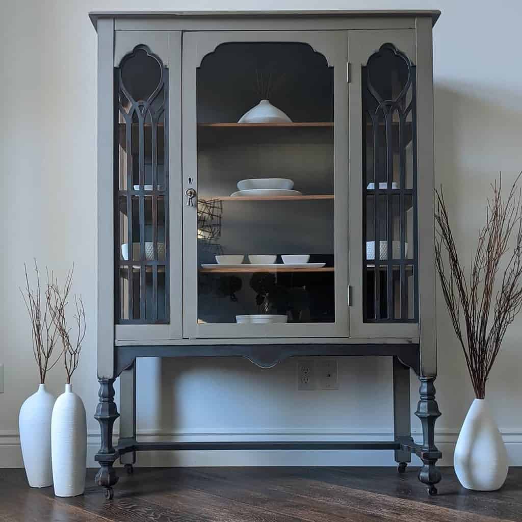 Elegant vintage china cabinet refinished in muted gray chalk paint with glass doors, intricate detailing, and a modern minimalist display